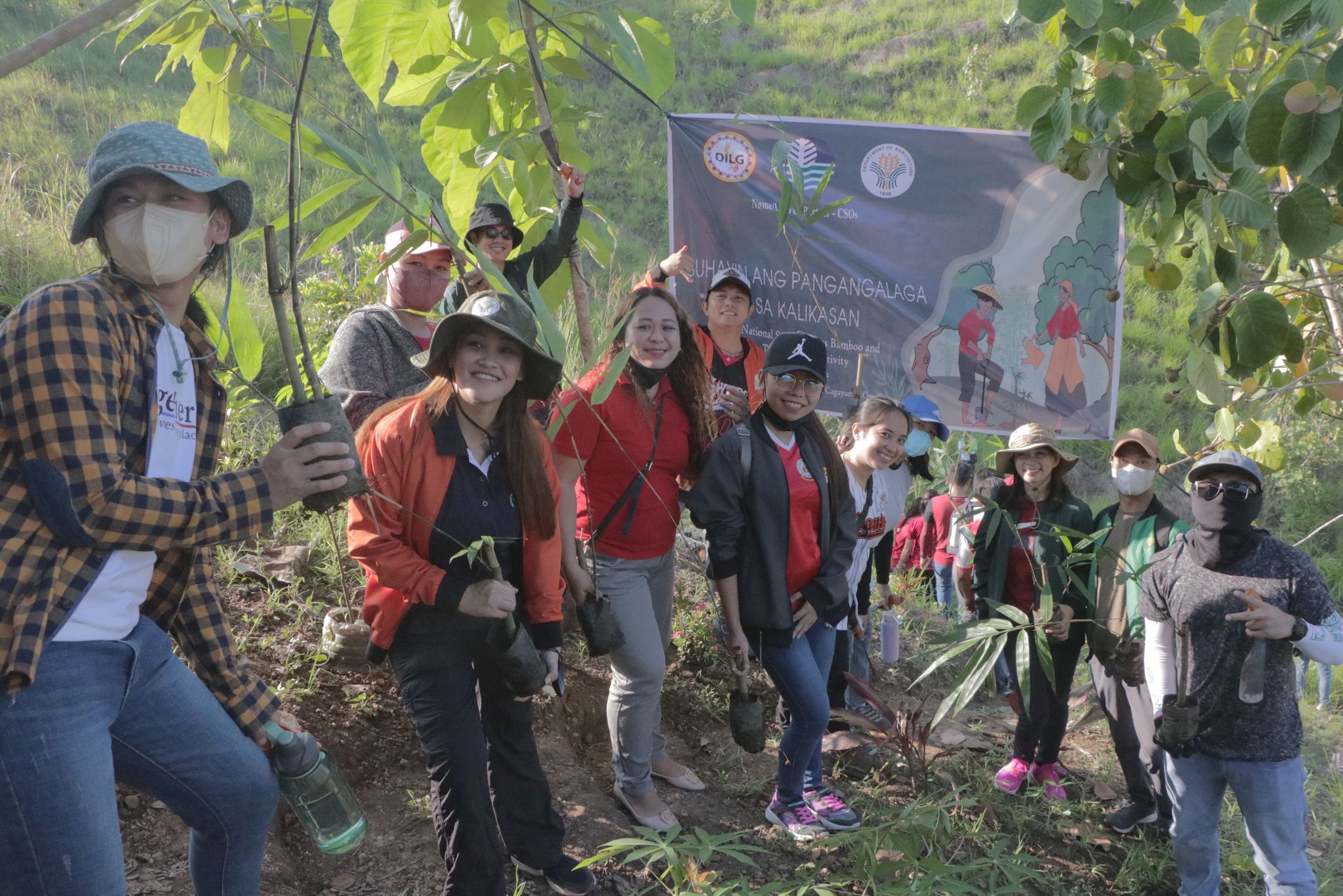 Volunteers holding seedlings beside a banner reading Suhayin ang Pangangalaga sa Kalikasan